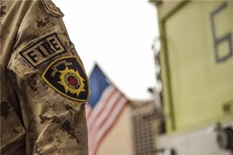 A Canadian Fire Fighter stands proudly beside an American fire truck  // Un pompier des Forces armées canadiennes se tient fièrement à côté d’un camion de pompiers américain.