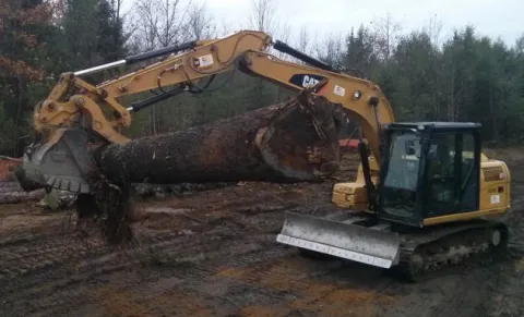 An excavator loading lumber onto a tractor trailer // Une excavatrice chargeant du bois à bord d’un camion gros porteur